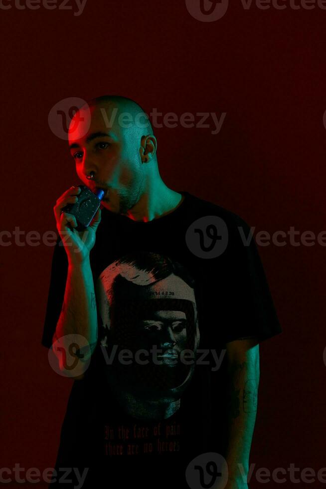 Close-up studio shot of a young bald guy vaping, blowing out a cloud of smoke on red background. photo