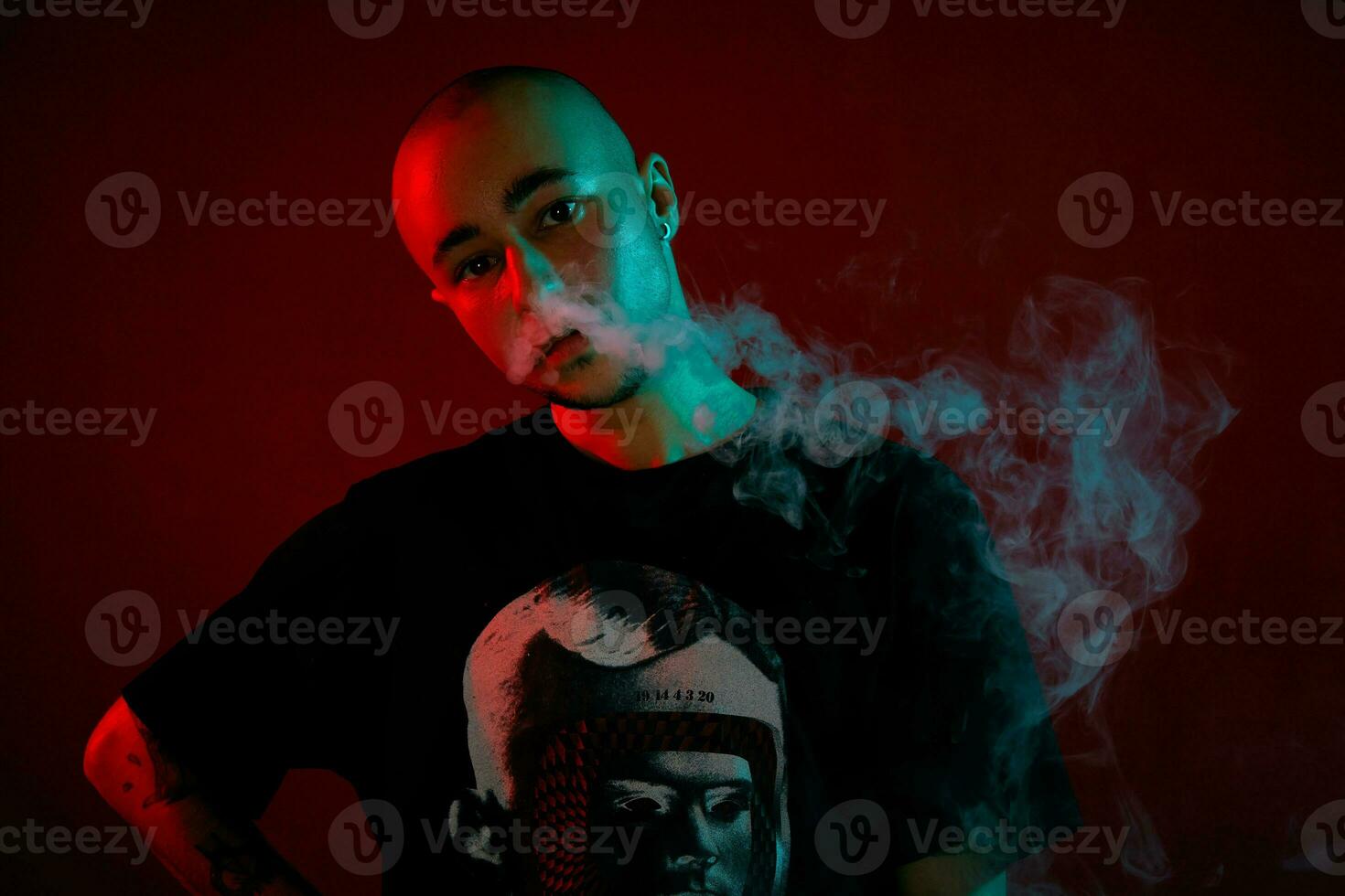 Close-up studio shot of a young bald guy vaping, blowing out a cloud of smoke on red background. photo