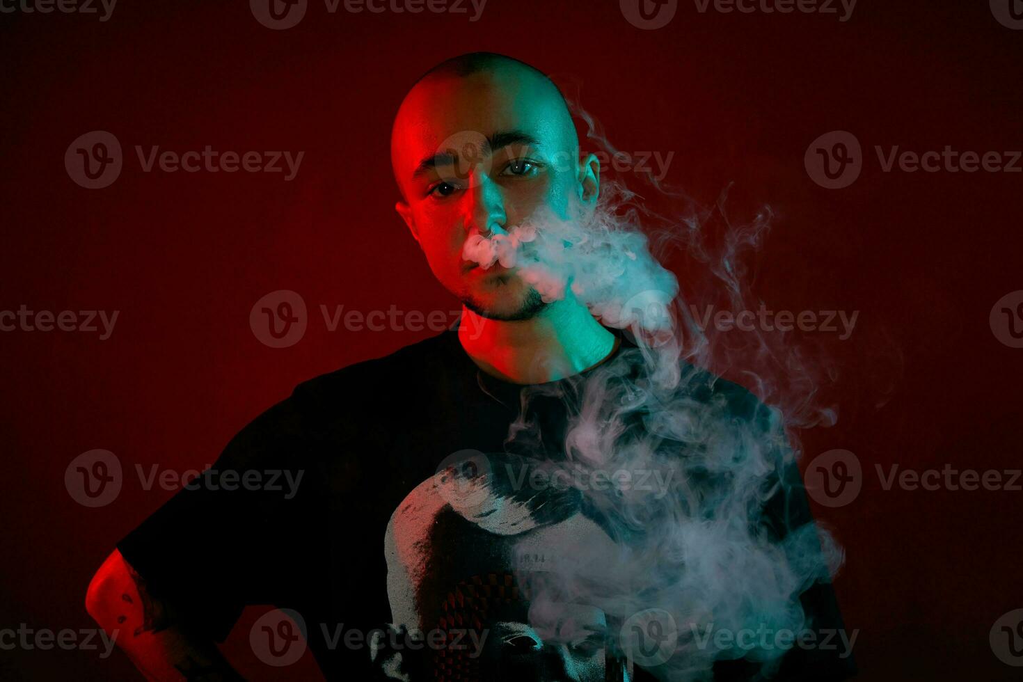 Close-up studio shot of a young bald guy vaping, blowing out a cloud of smoke on red background. photo