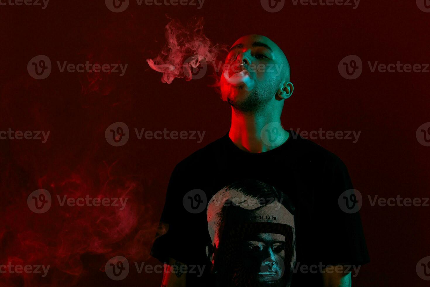 Close-up studio shot of a young bald guy vaping, blowing out a cloud of smoke on red background. photo