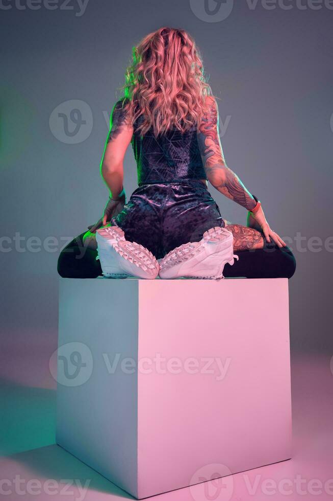 Young fitness blonde woman wearing blue velour booty shorts posing on camera, sits on top of big cube, back view photo