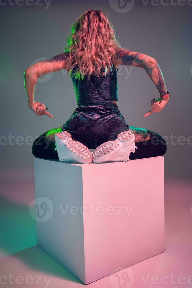 Young fitness blonde woman wearing blue velour booty shorts posing on camera, sits on top of big cube, back view photo