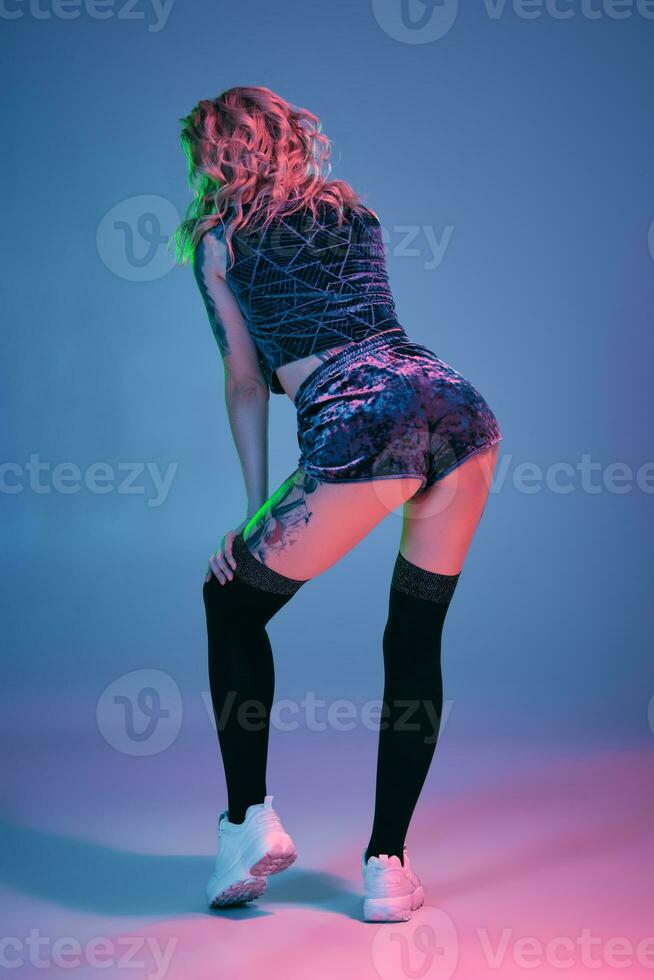 Young blonde woman in velour blue sport wear and black stockings seductively posing on camera in studio. Pink and blue background photo