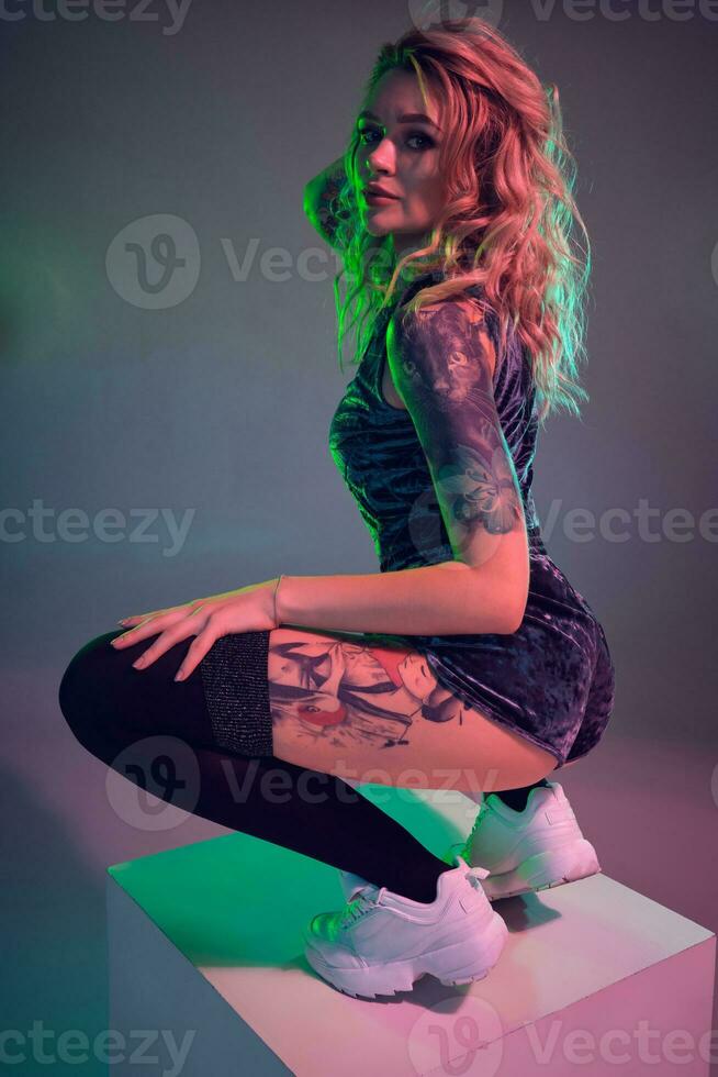 Young fitness blonde woman wearing blue velour booty shorts posing on camera, making squats on top of big cube photo