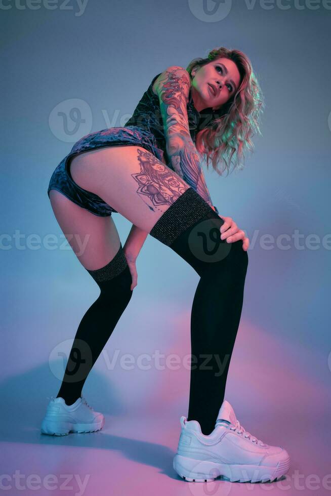 Young blonde woman in velour blue sport wear and black stockings seductively posing on camera in studio. Pink and blue background photo