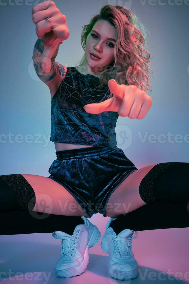 Young fitness blonde woman wearing blue velour booty shorts squats in front of camera, looking at camera, pink and blue background photo