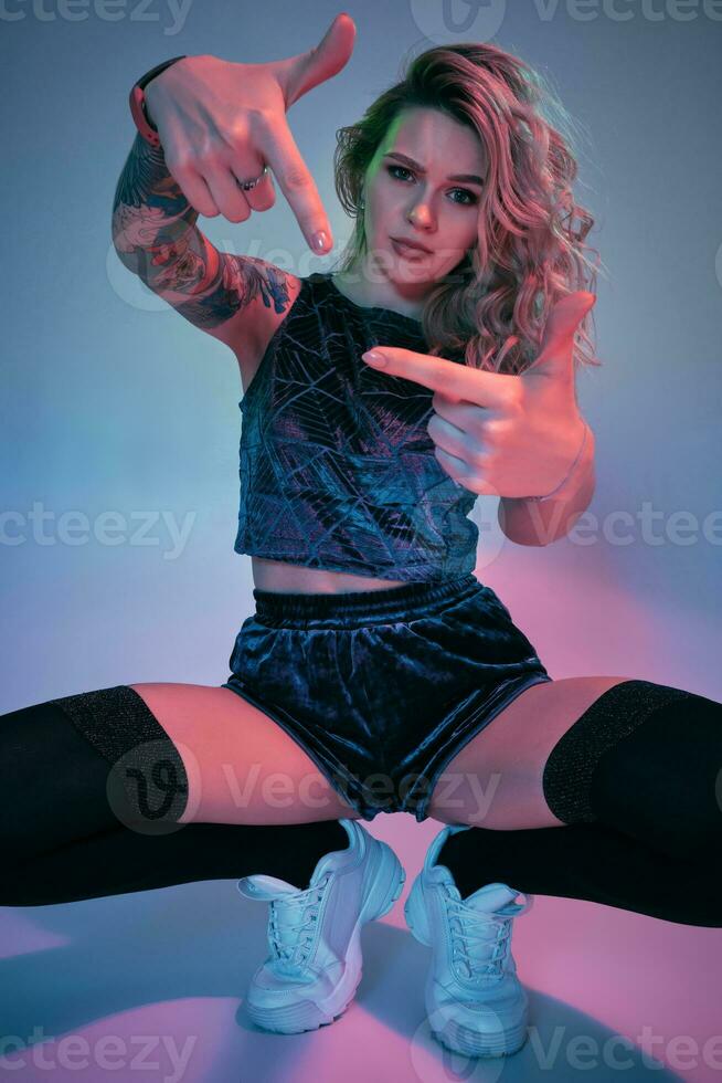 Young fitness blonde woman wearing blue velour booty shorts squats in front of camera, looking at camera, pink and blue background photo