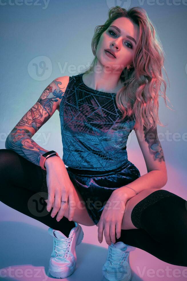 Young fitness blonde woman wearing blue velour booty shorts squats in front of camera, looking at camera, pink and blue background photo