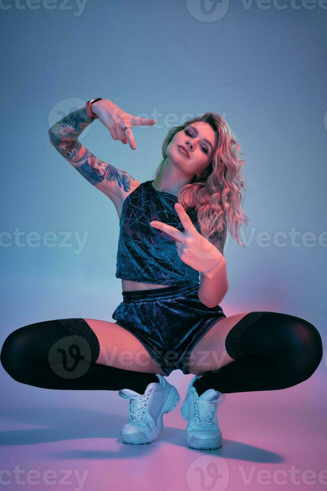 Young fitness blonde woman wearing blue velour booty shorts squats in front of camera, looking at camera, pink and blue background photo