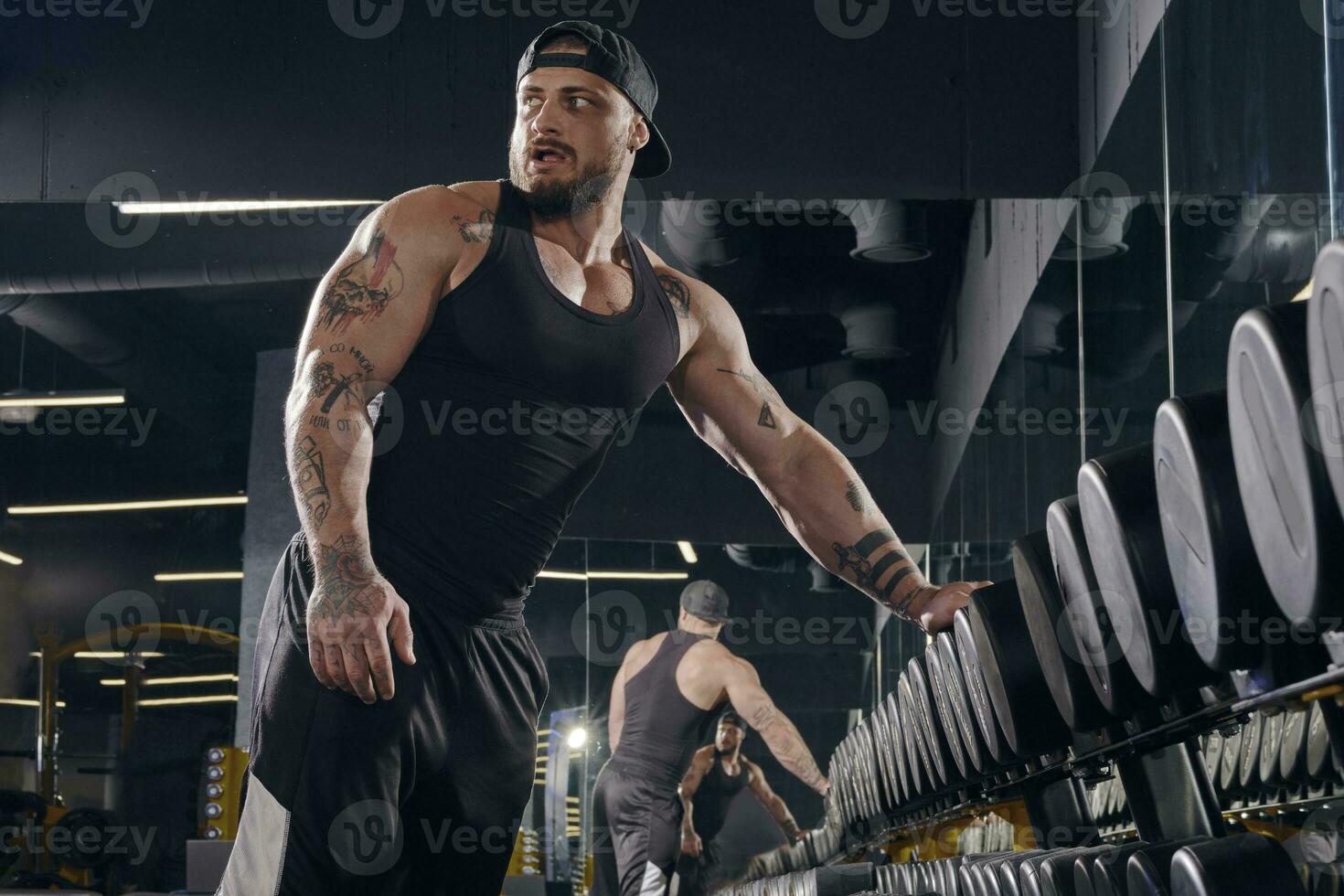 Young, tattooed male in black shorts, vest, cap. Going to do exercises with dumbbells for training his muscles in dark gym. Sport, fitness. Close up photo