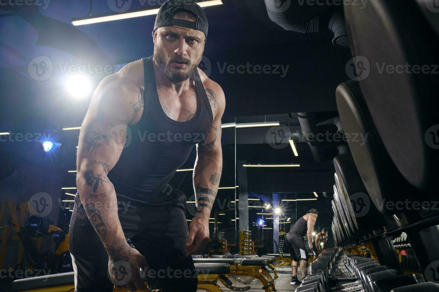 elegante, tatuado, barbado deportista en negro bermudas, chaleco, gorra. mirando a usted mientras en pie cerca conjunto de negro mancuernas oscuro gimnasia. cerca arriba foto