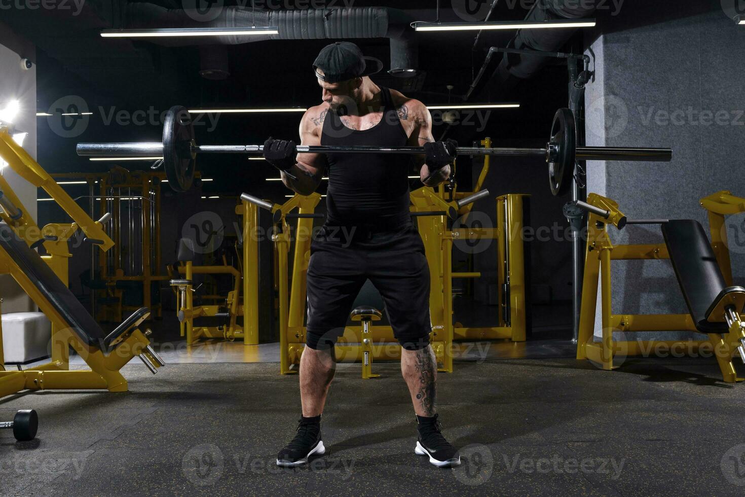 Bearded sportsman in black sport gloves, shorts, vest, cap and sneakers. He lifting a barbell, posing in dark gym with yellow equipment. Full length photo