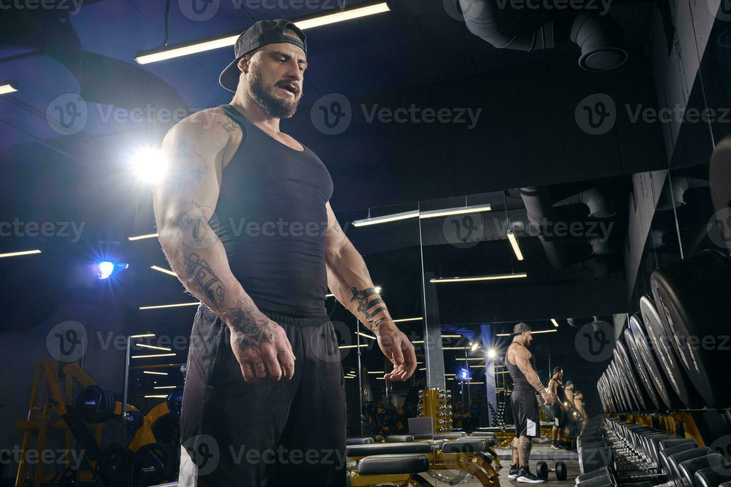 Tattooed, bearded athlete in black shorts, vest, cap. Going to do exercises with dumbbells, looking at set of black weights. Dark gym. Close up photo