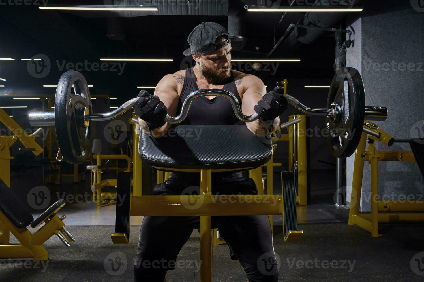 Tattooed athlete in black sport gloves, shorts, vest, cap. Lifting barbell, training his biceps, sitting on preacher curl bench at dark gym. Close up photo