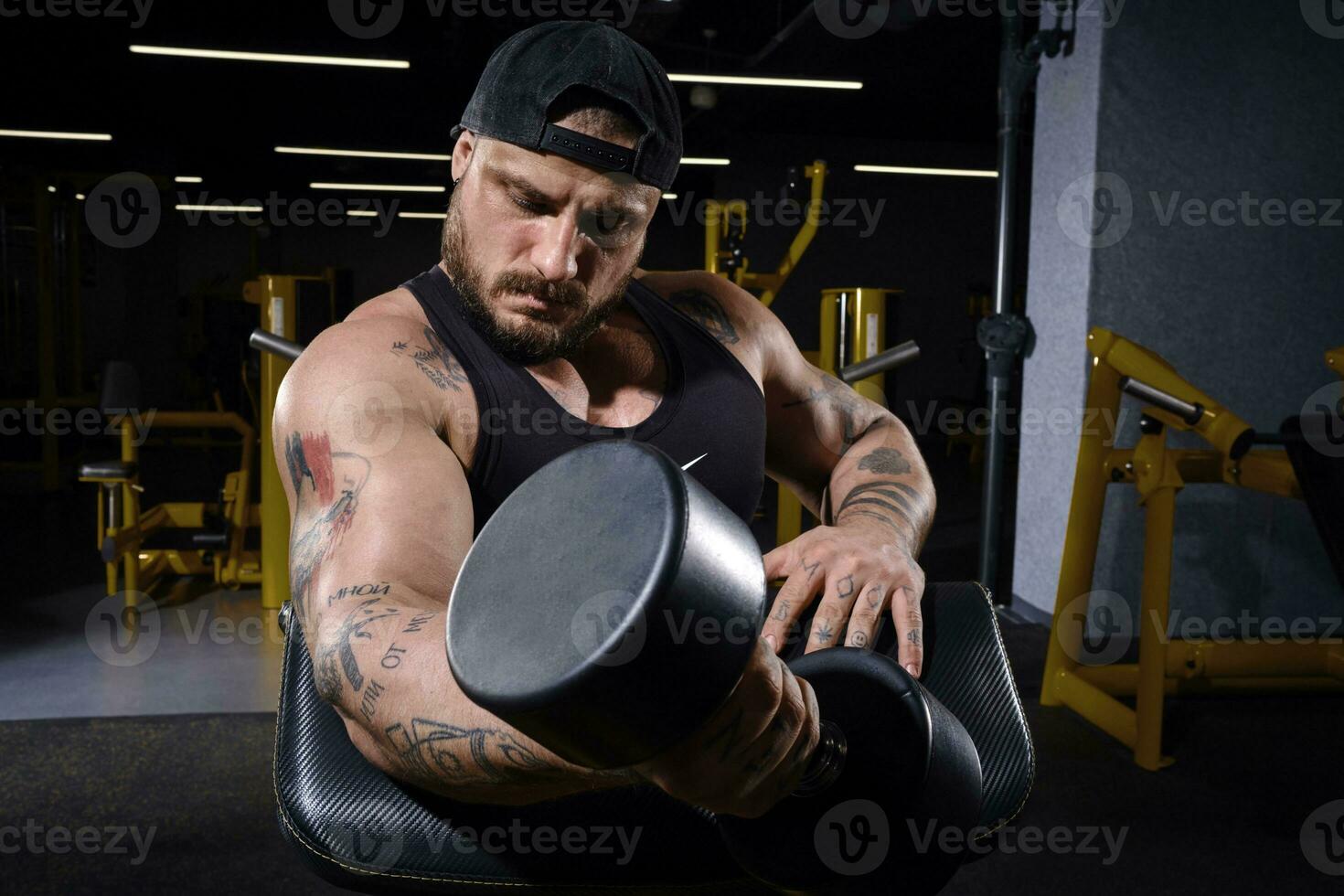 tatuado, barbado chico en negro chaleco y gorra. él es levantamiento un pesa, formación su bíceps, sentado en predicador rizo banco a oscuro gimnasia. cerca arriba foto