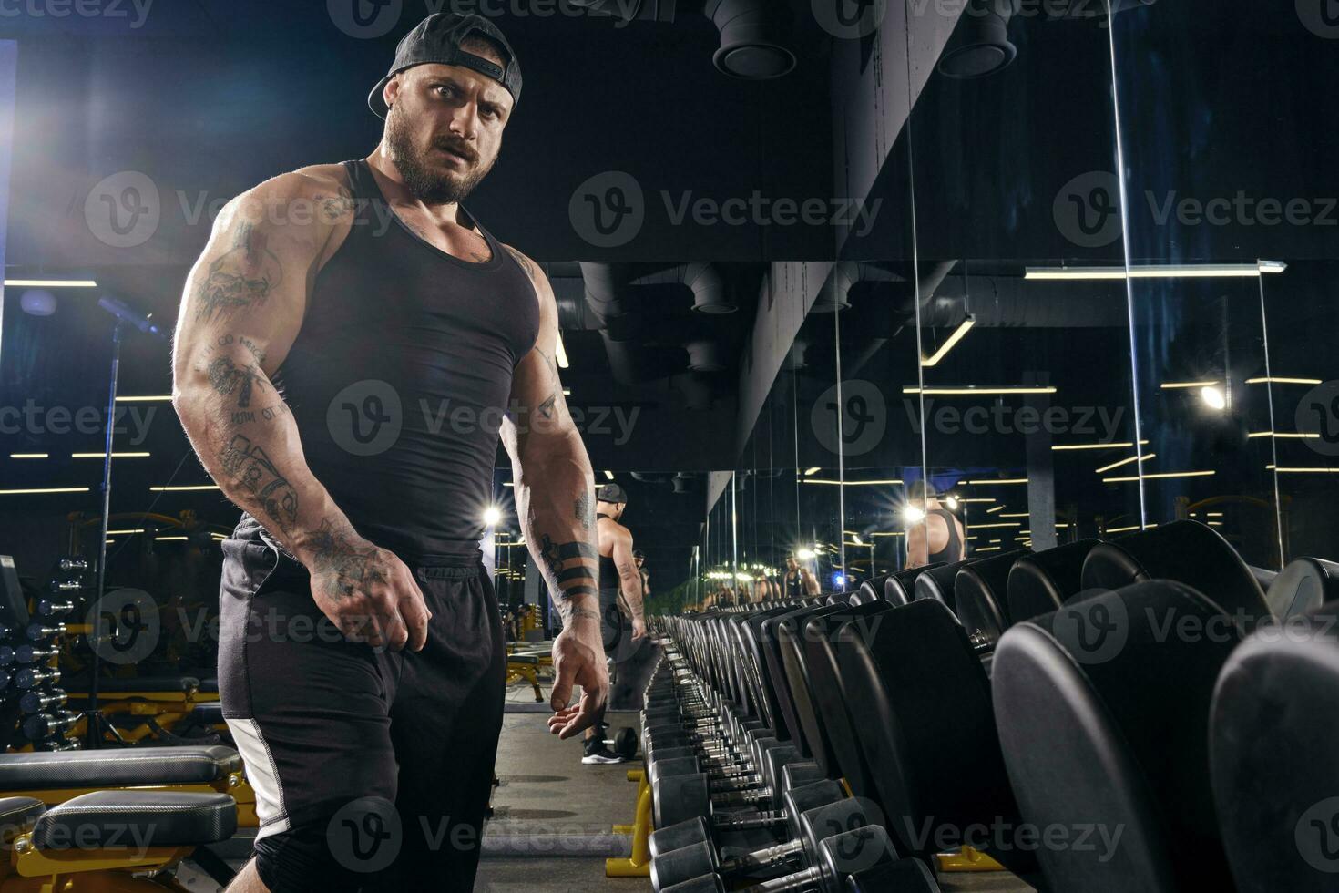 Attractive, tattooed, bearded man in black shorts, vest, cap. Looking at you, standing near set of black dumbbells. Dark gym. Close up photo