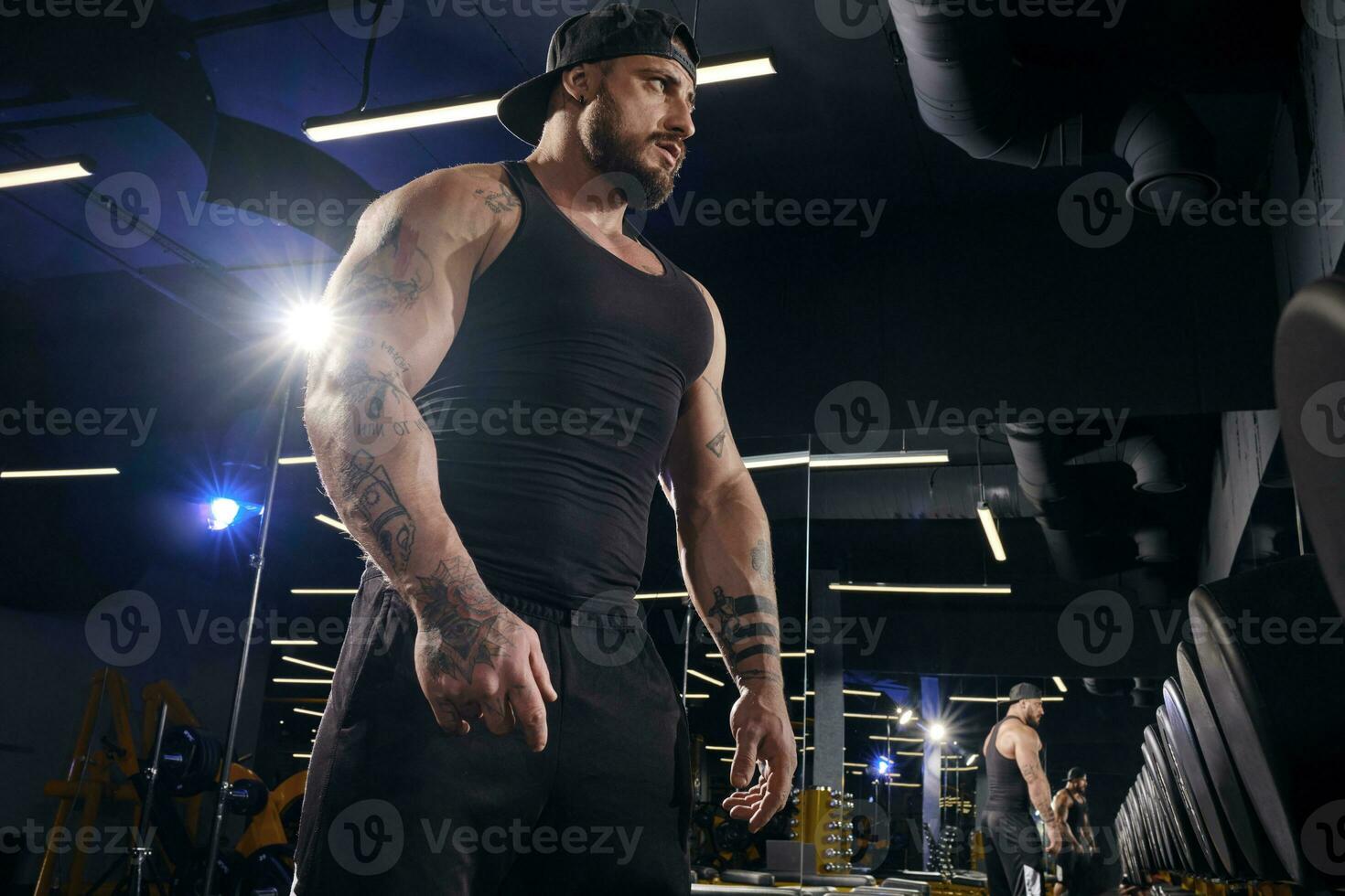 Tattooed male in black shorts, vest, cap. Going to do exercises with dumbbells, looking at mirror near set of black weights. Dark gym. Close up photo