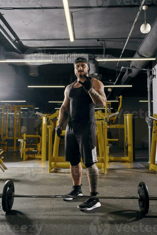 Attractive, tattooed, bearded man in black sport gloves, shorts, vest, cap and sneakers. Showing cool sign, barbell is on floor. Dark gym. Full length photo