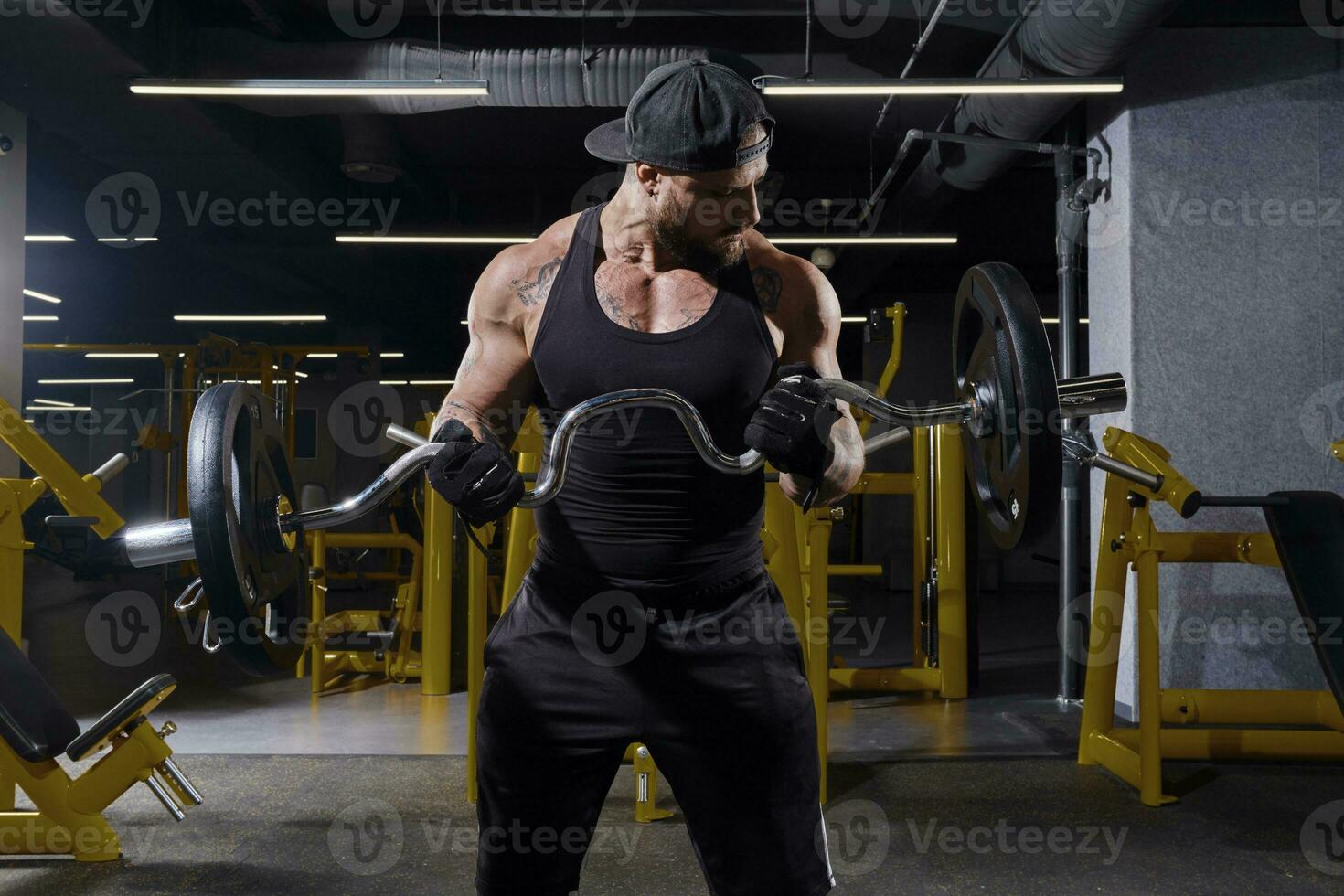 Sportsman in black sport gloves, shorts, vest and cap. He is lifting barbell, training his biceps, posing in dark gym with yellow equipment. Close up photo