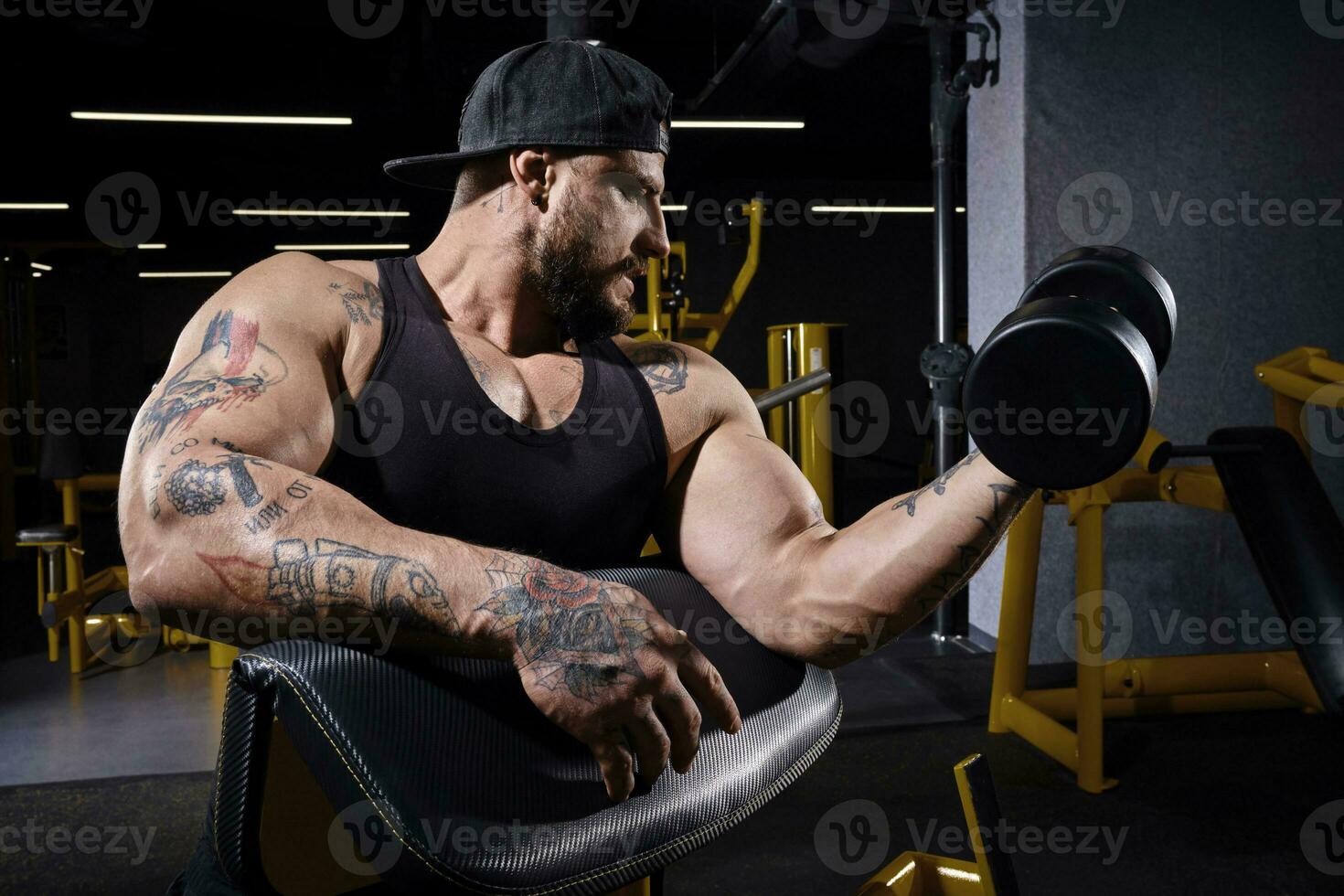 tatuado chico con fuerte cuerpo, en negro chaleco y gorra. él levantamiento pesa, formación su bíceps, sentado en predicador rizo banco a oscuro gimnasia. cerca arriba foto