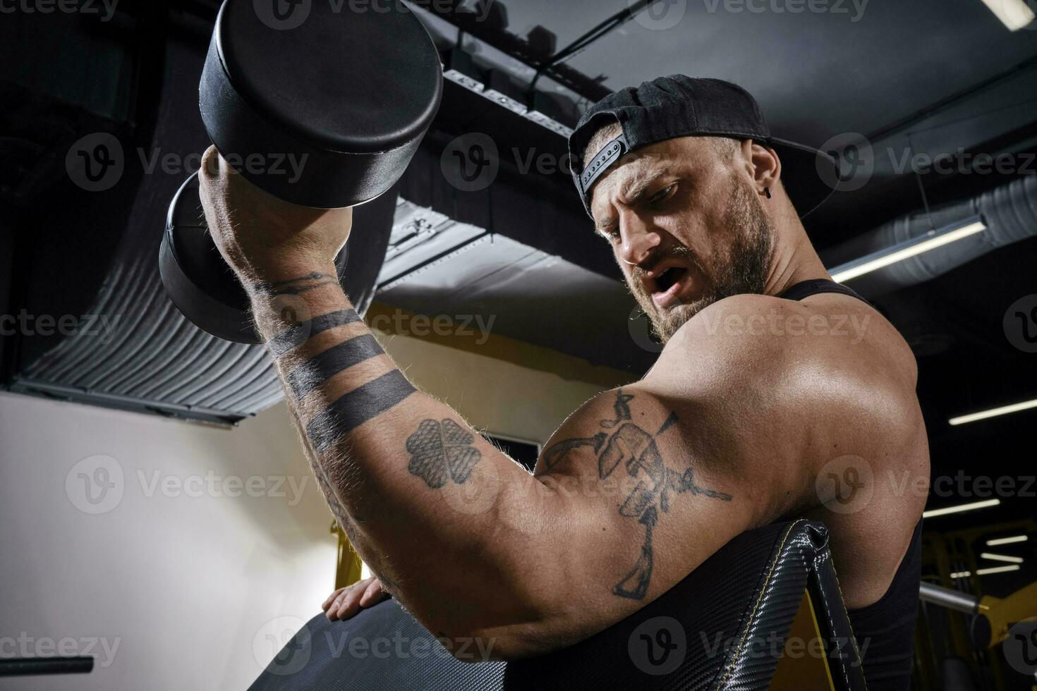 Tattooed, bearded man in black vest and cap. He is lifting a dumbbell, training his biceps, sitting on preacher curl bench at dark gym. Close up photo