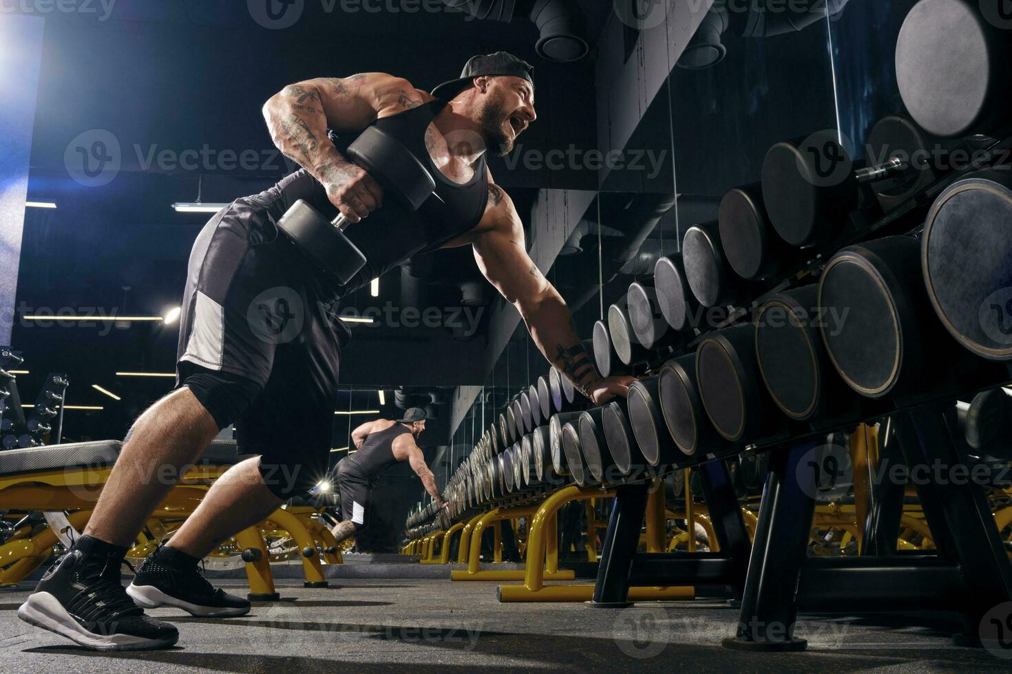 Muscular male in black shorts, vest, cap. Exercising with dumbbells for training his triceps, leaning on set of black weights. Dark gym. Close up photo