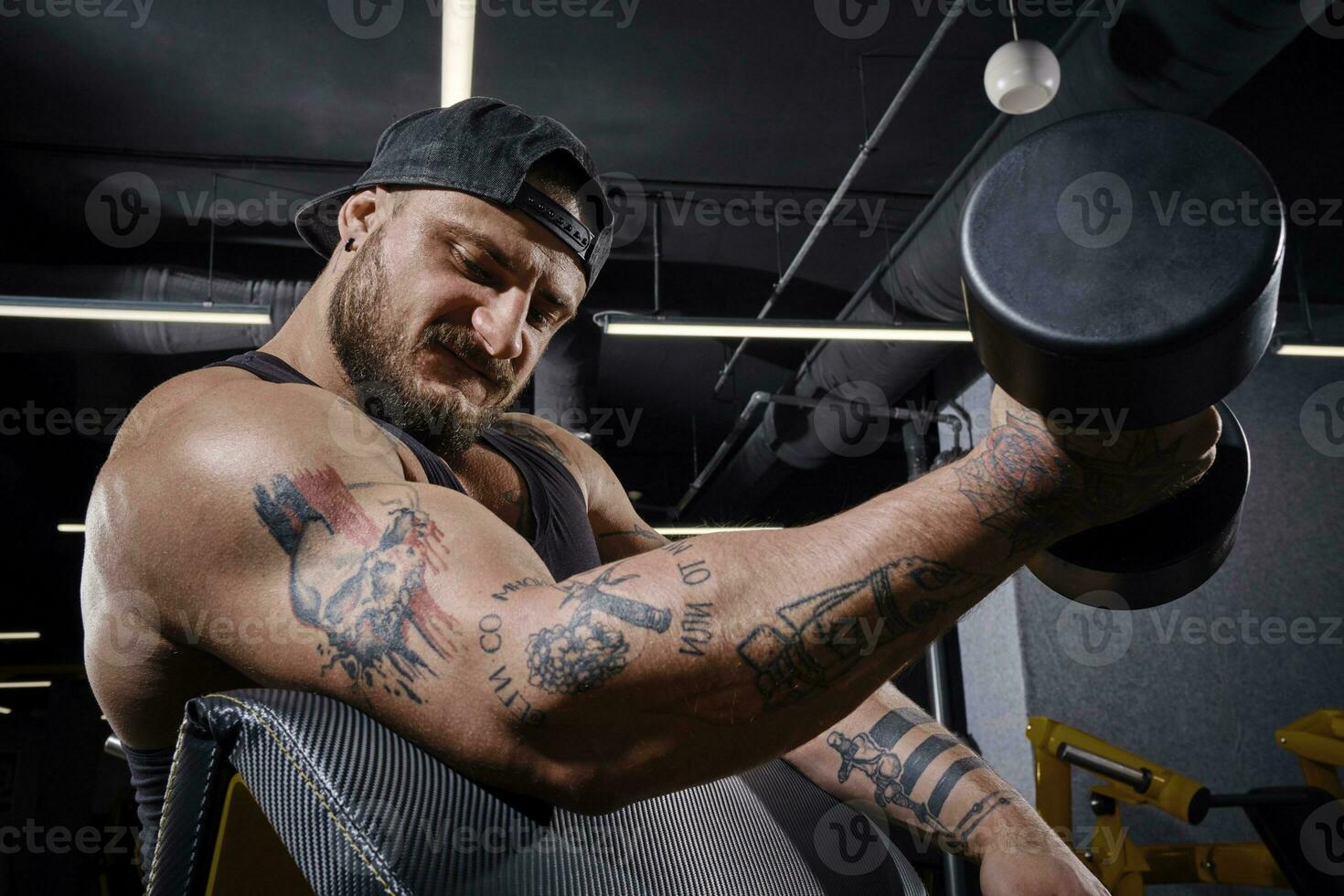 Tattooed, bearded male in black vest and cap. He is lifting a dumbbell, training his biceps, sitting on preacher curl bench at dark gym. Close up photo