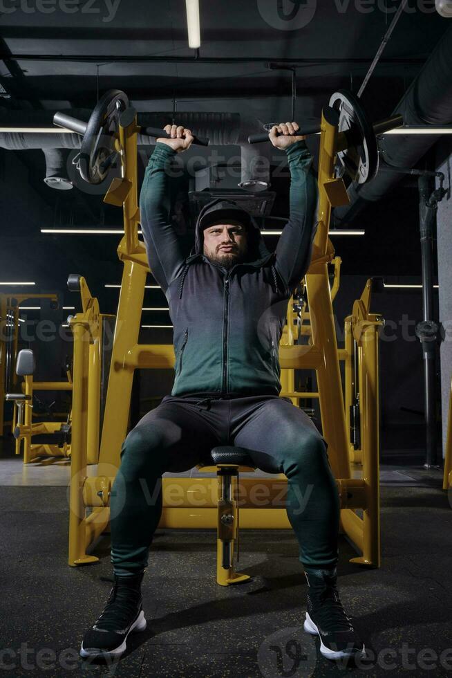 masculino en negro chandal con un capucha, gorra y zapatillas. él ejecutando cofre prensa, sentado en un ejercicio máquina, posando en oscuro gimnasia. lleno longitud foto