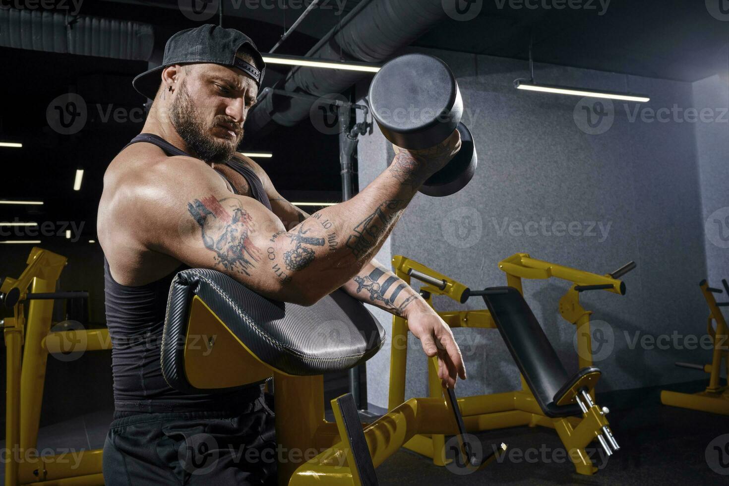 barbado atleta en negro chaleco y gorra. él es levantamiento un pesa, formación su bíceps, sentado en predicador rizo banco a oscuro gimnasia. cerca arriba foto