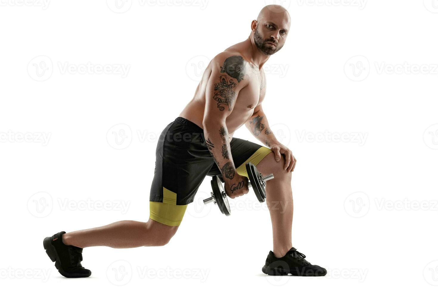 Athletic bald, tattooed man in black shorts and sneakers is posing with a dumbbell isolated on white background. Full length portrait. photo