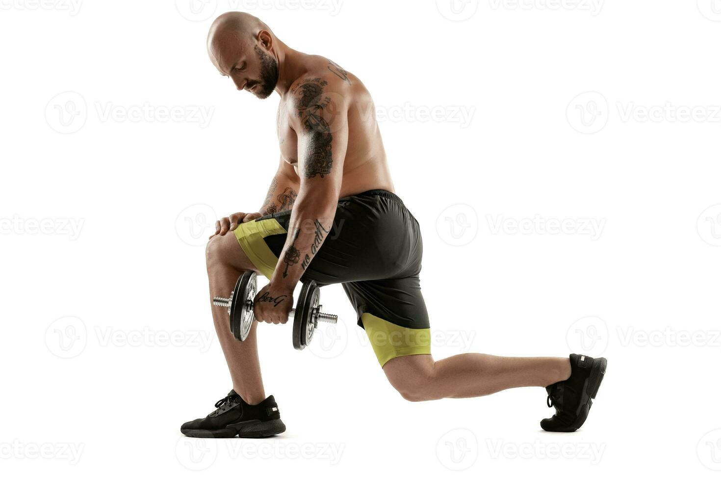 Athletic bald, tattooed man in black shorts and sneakers is posing with a dumbbell isolated on white background. Full length portrait. photo