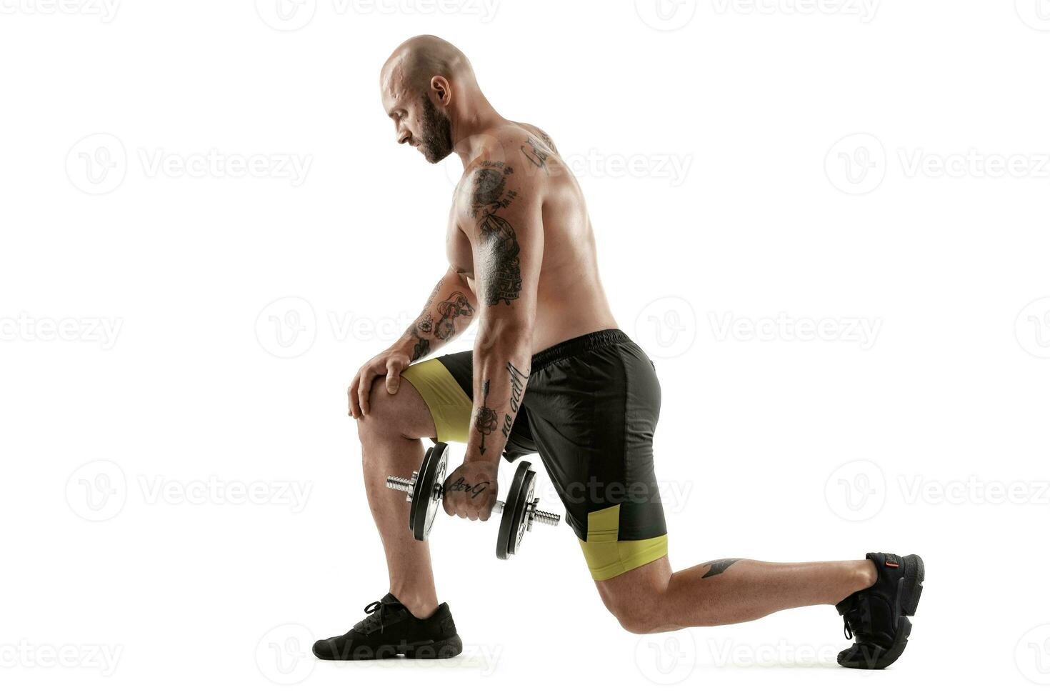 Athletic bald, tattooed man in black shorts and sneakers is posing with a dumbbell isolated on white background. Full length portrait. photo