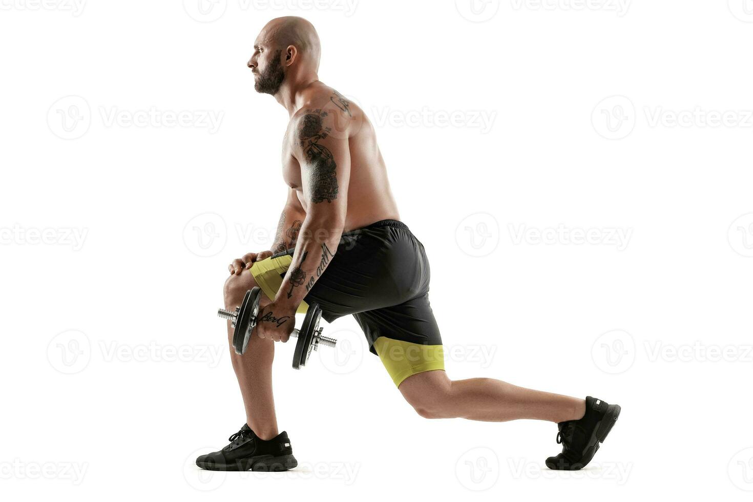 Athletic bald, tattooed man in black shorts and sneakers is posing with a dumbbell isolated on white background. Full length portrait. photo