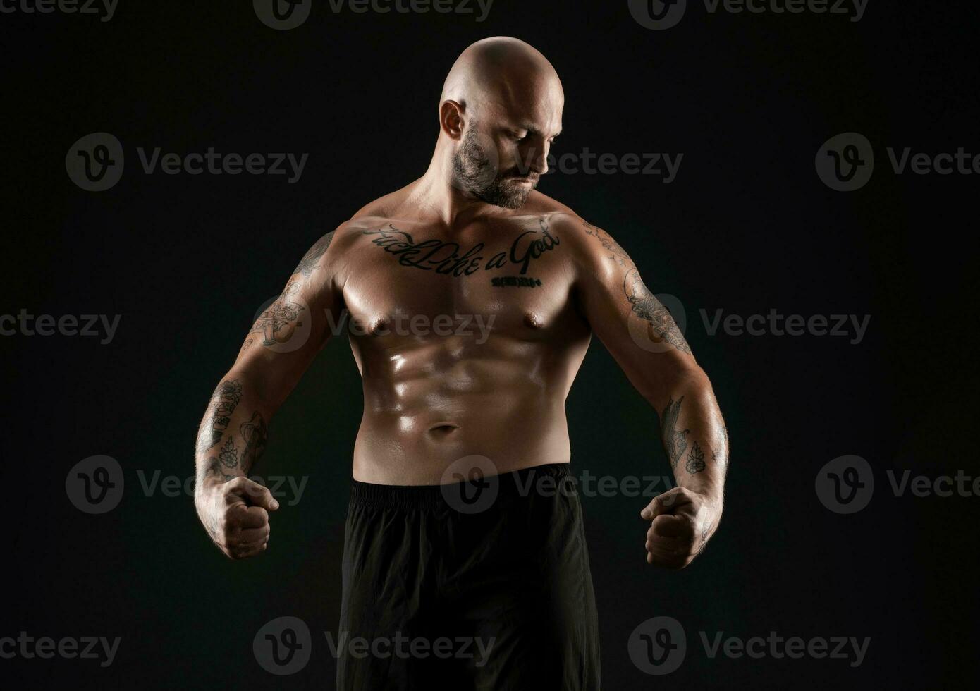 Athletic bald, bearded, tattooed man in black shorts is posing against a black background. Close-up portrait. photo