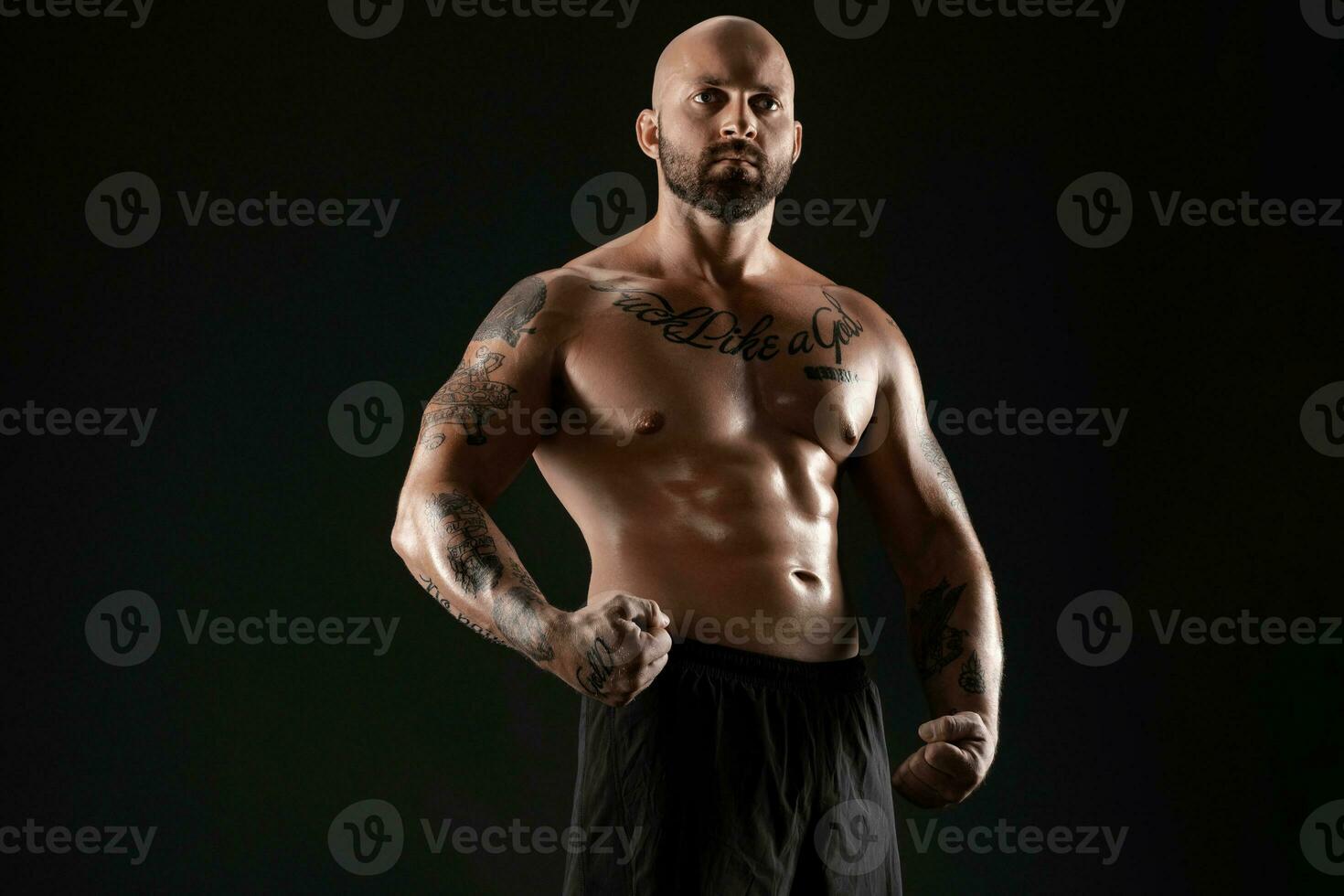 atlético calvo, barbado, tatuado hombre en negro pantalones cortos es posando en contra un negro antecedentes. de cerca retrato. foto