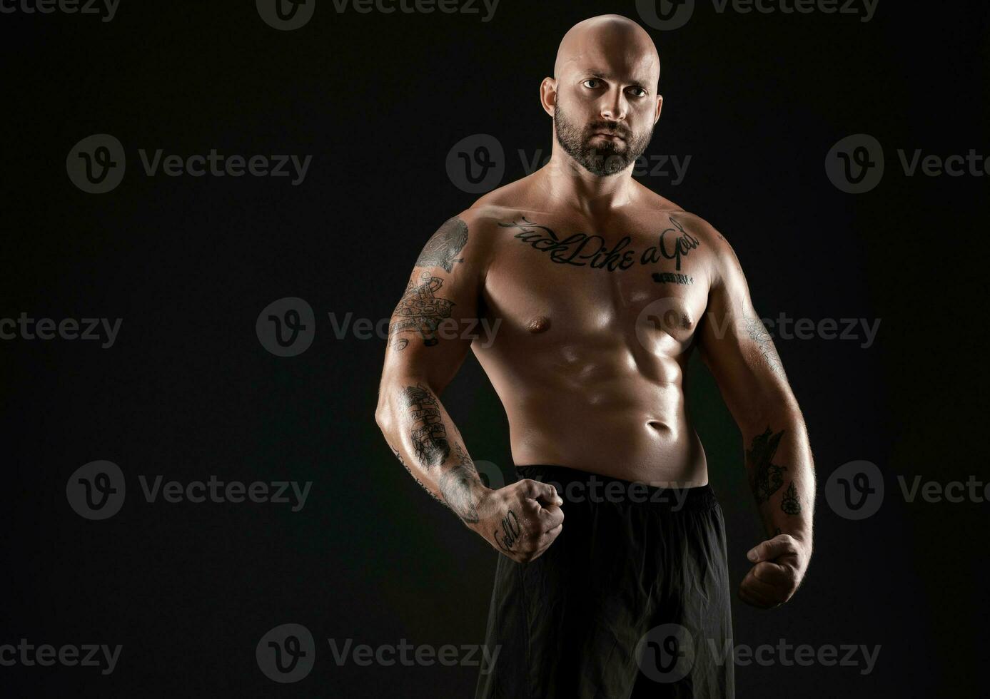 Athletic bald, bearded, tattooed man in black shorts is posing against a black background. Close-up portrait. photo