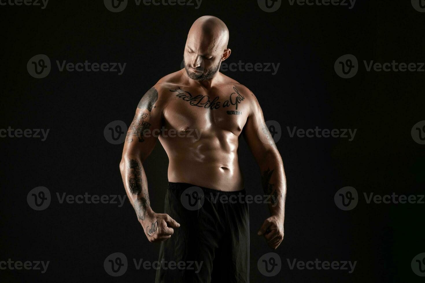 Athletic bald, bearded, tattooed man in black shorts is posing against a black background. Close-up portrait. photo