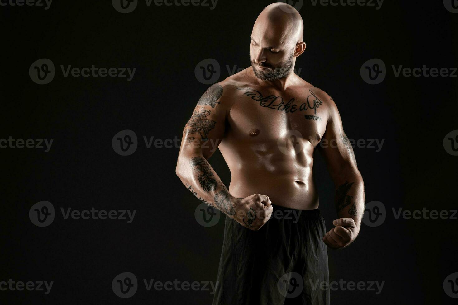 Athletic bald, bearded, tattooed man in black shorts is posing against a black background. Close-up portrait. photo