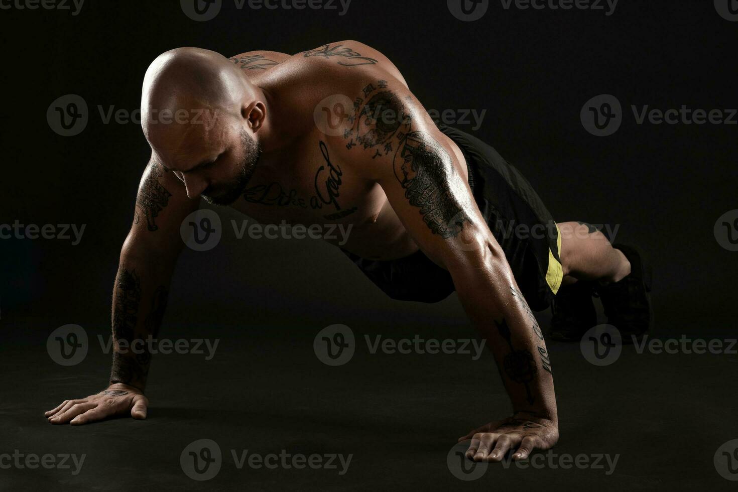 atlético calvo, tatuado hombre en negro pantalones cortos y zapatillas es posando en contra un negro antecedentes. de cerca retrato. foto