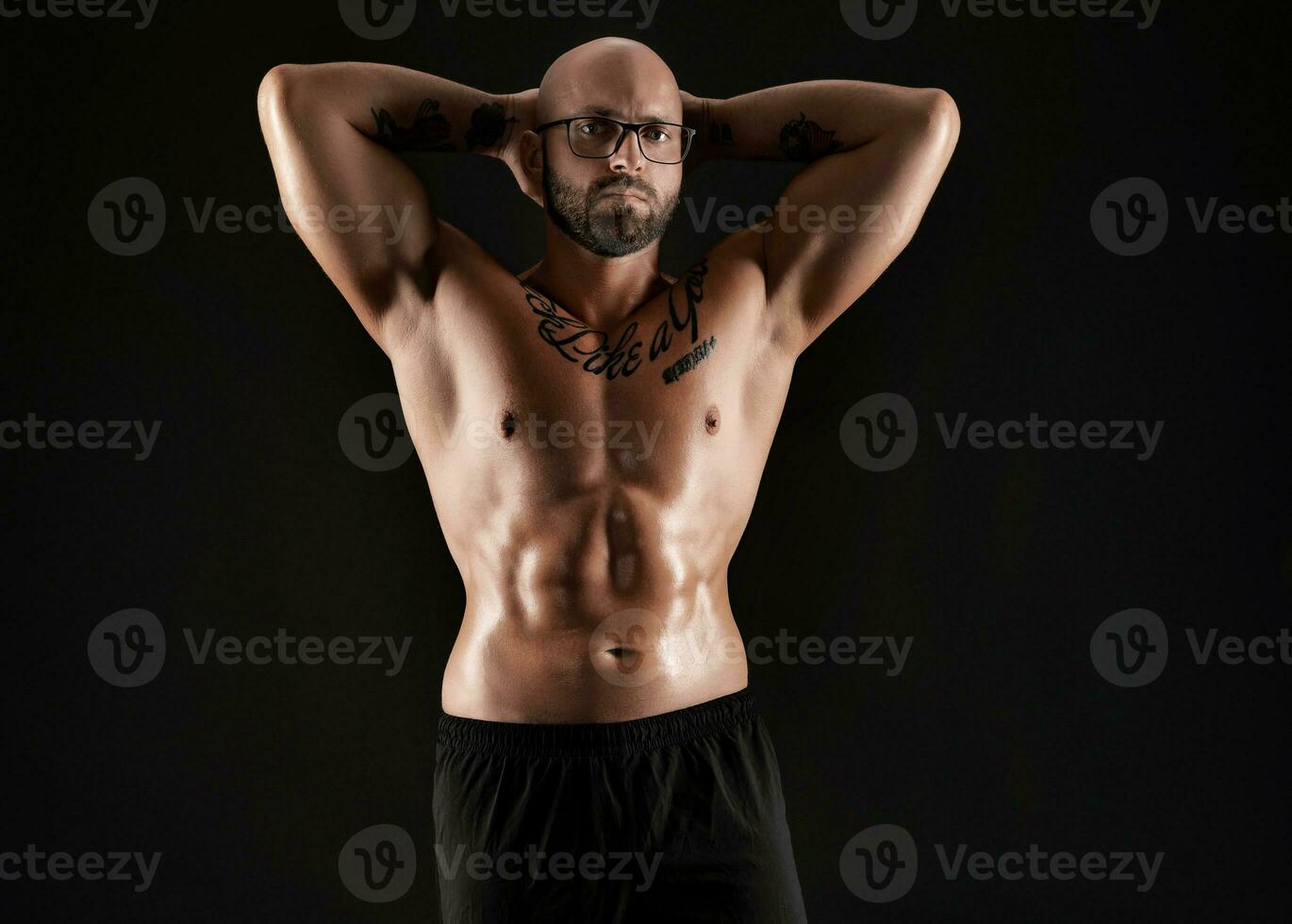 atlético calvo, barbado, tatuado hombre en negro pantalones cortos es posando en contra un negro antecedentes. de cerca retrato. foto
