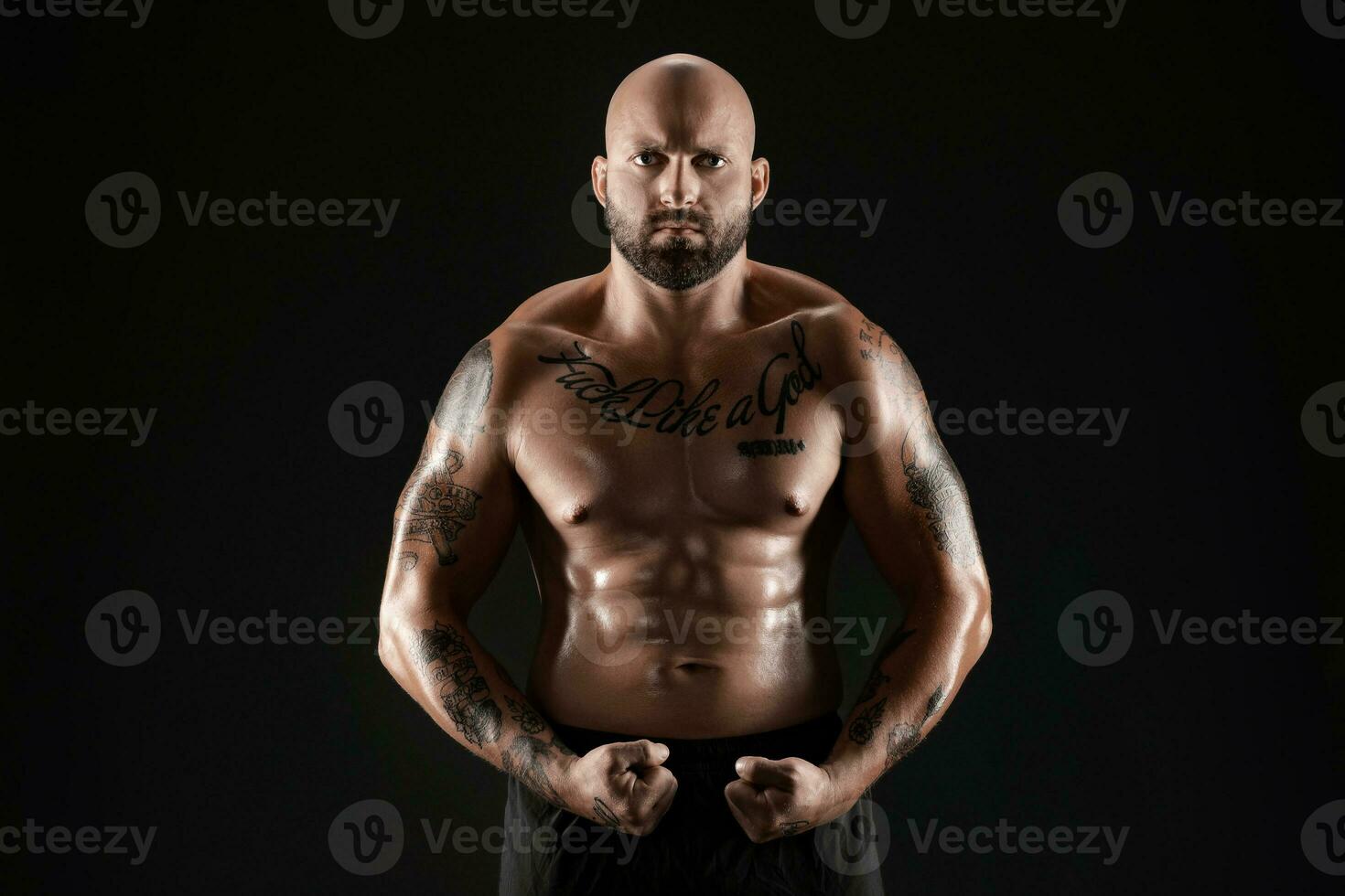 Athletic bald, bearded, tattooed man in black shorts is posing against a black background. Close-up portrait. photo