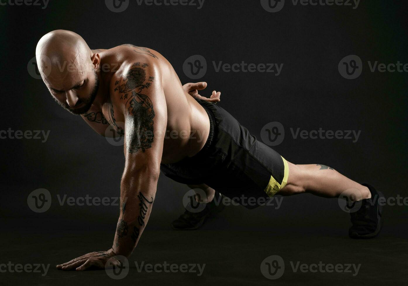 atlético calvo, tatuado hombre en negro pantalones cortos y zapatillas es posando en contra un negro antecedentes. de cerca retrato. foto