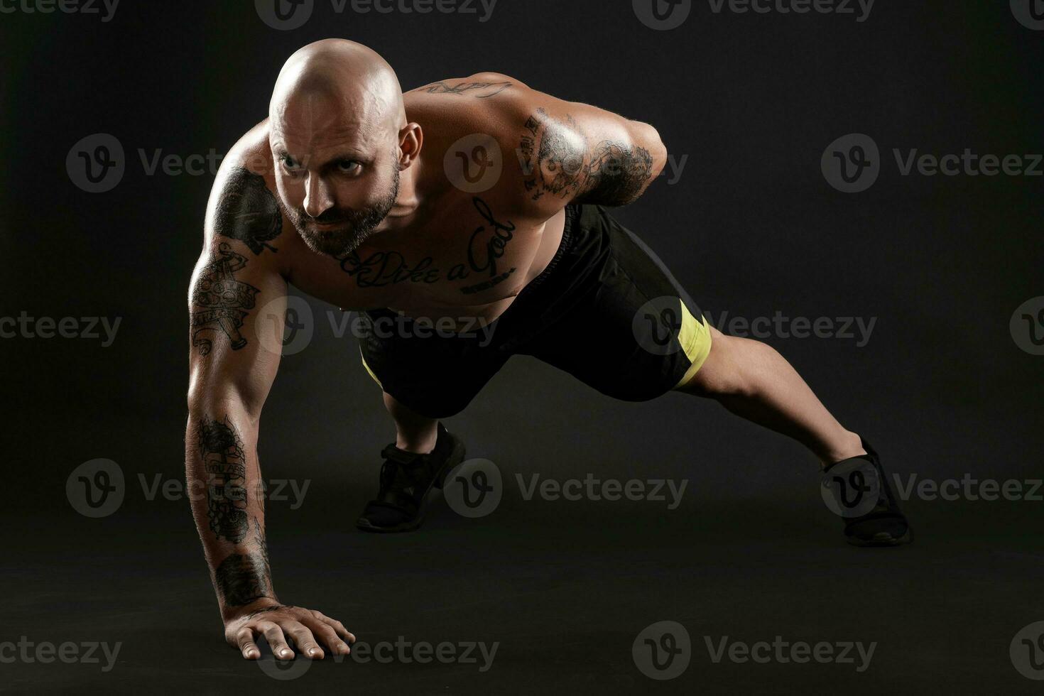 Athletic bald, tattooed man in black shorts and sneakers is posing against a black background. Close-up portrait. photo