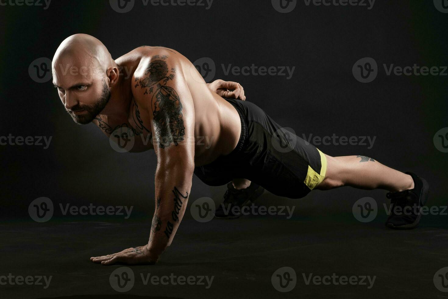 atlético calvo, tatuado hombre en negro pantalones cortos y zapatillas es posando en contra un negro antecedentes. de cerca retrato. foto