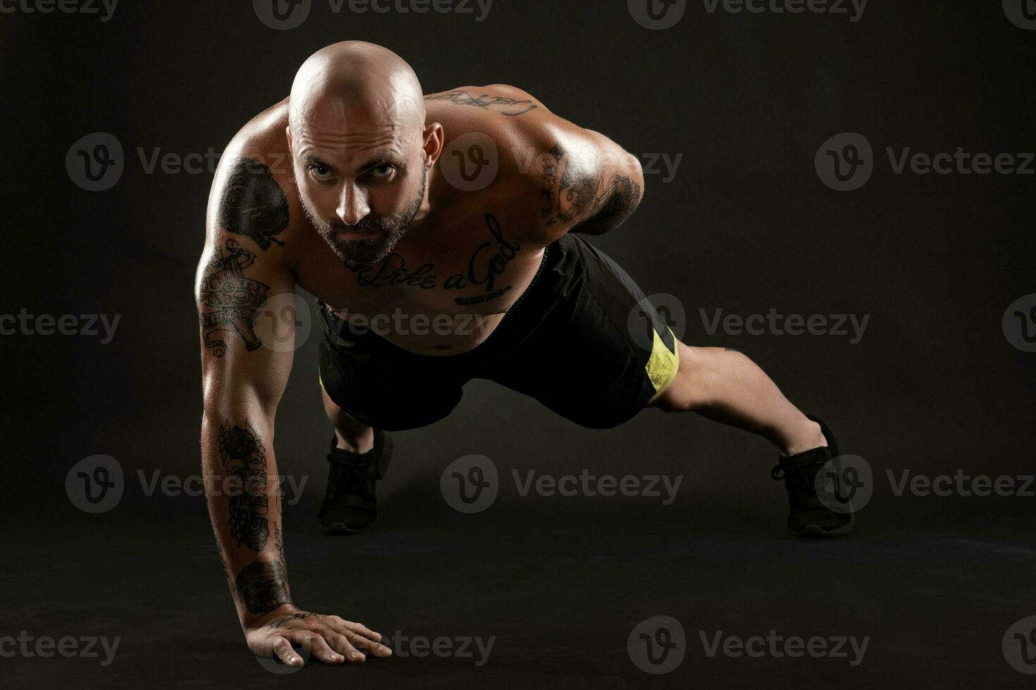 Athletic bald, tattooed man in black shorts and sneakers is posing against a black background. Close-up portrait. photo