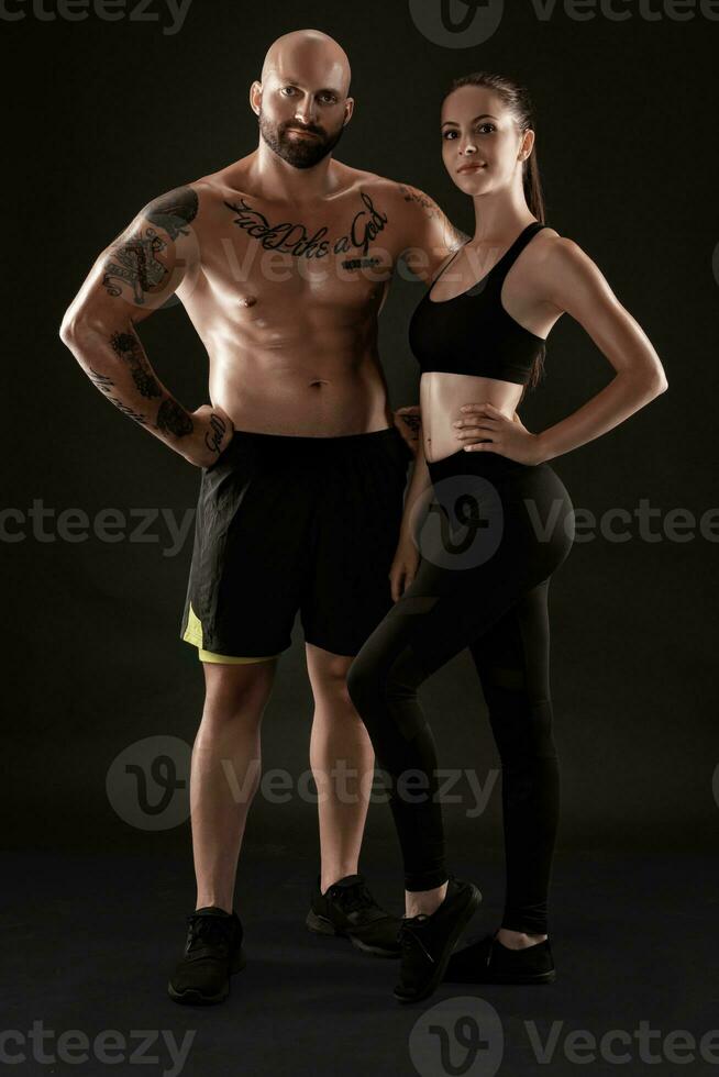 Athletic man in shorts and sneakers with brunette woman in leggings and top posing on black background. Fitness couple, gym concept. photo