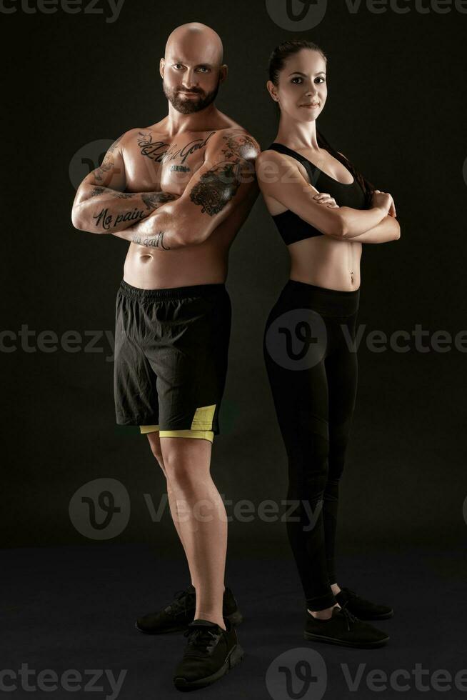 Athletic man in shorts and sneakers with brunette woman in leggings and top posing on black background. Fitness couple, gym concept. photo