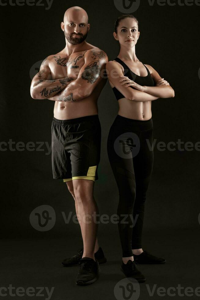 Athletic man in shorts and sneakers with brunette woman in leggings and top posing on black background. Fitness couple, gym concept. photo