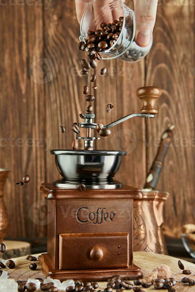 Pours coffee beans into vintage coffee grinder, coffee brewer and copper cup on wooden background photo