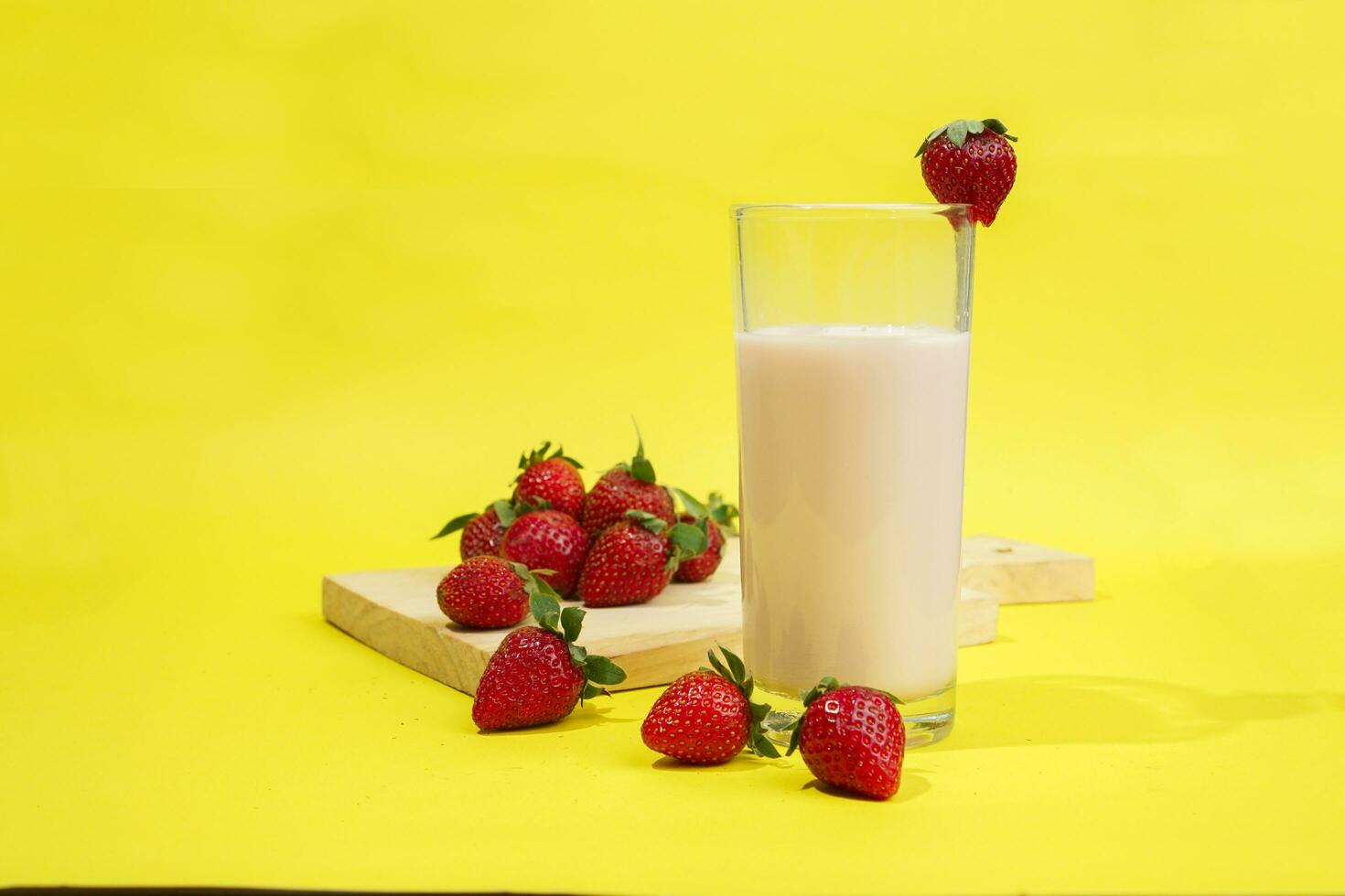 a glass of milk with strawberry garnish on cut board isolated yellow background photo